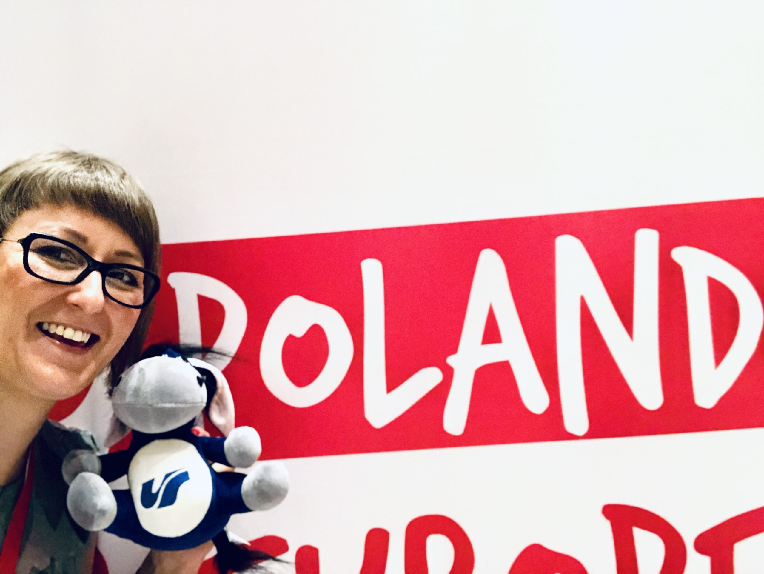 Po lewej uśmiechnięta dziewczyna w okularach, w ręce trzyma maskotkę: osła z logo Uniwersytetu Śląskiego na brzuchu. W tle napis: Poland