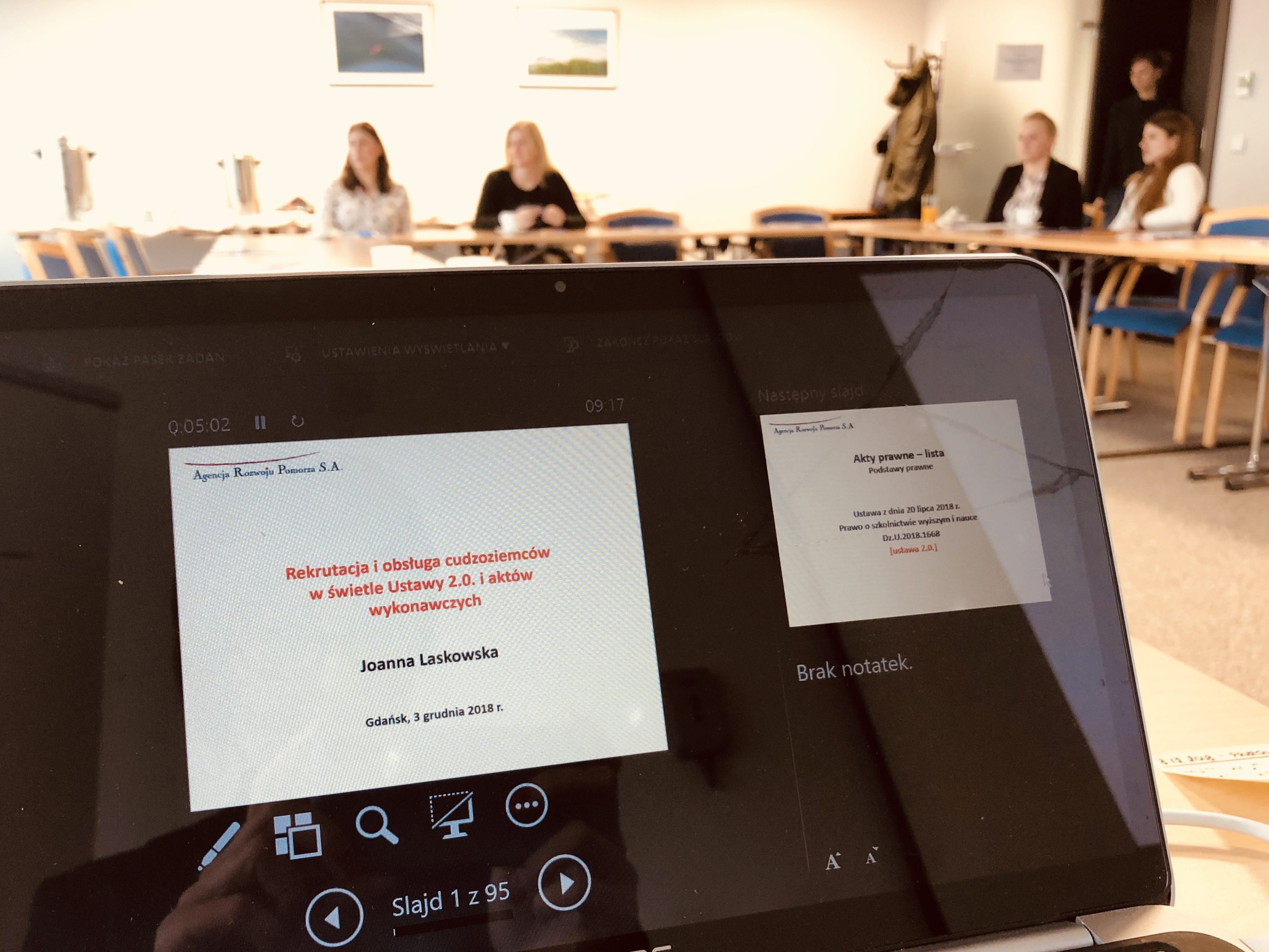 Pierwszy plan: laptop z otwartą prezentacją na temat rekrutacji cudzoziemców. W tle ludzie w okół stołu.