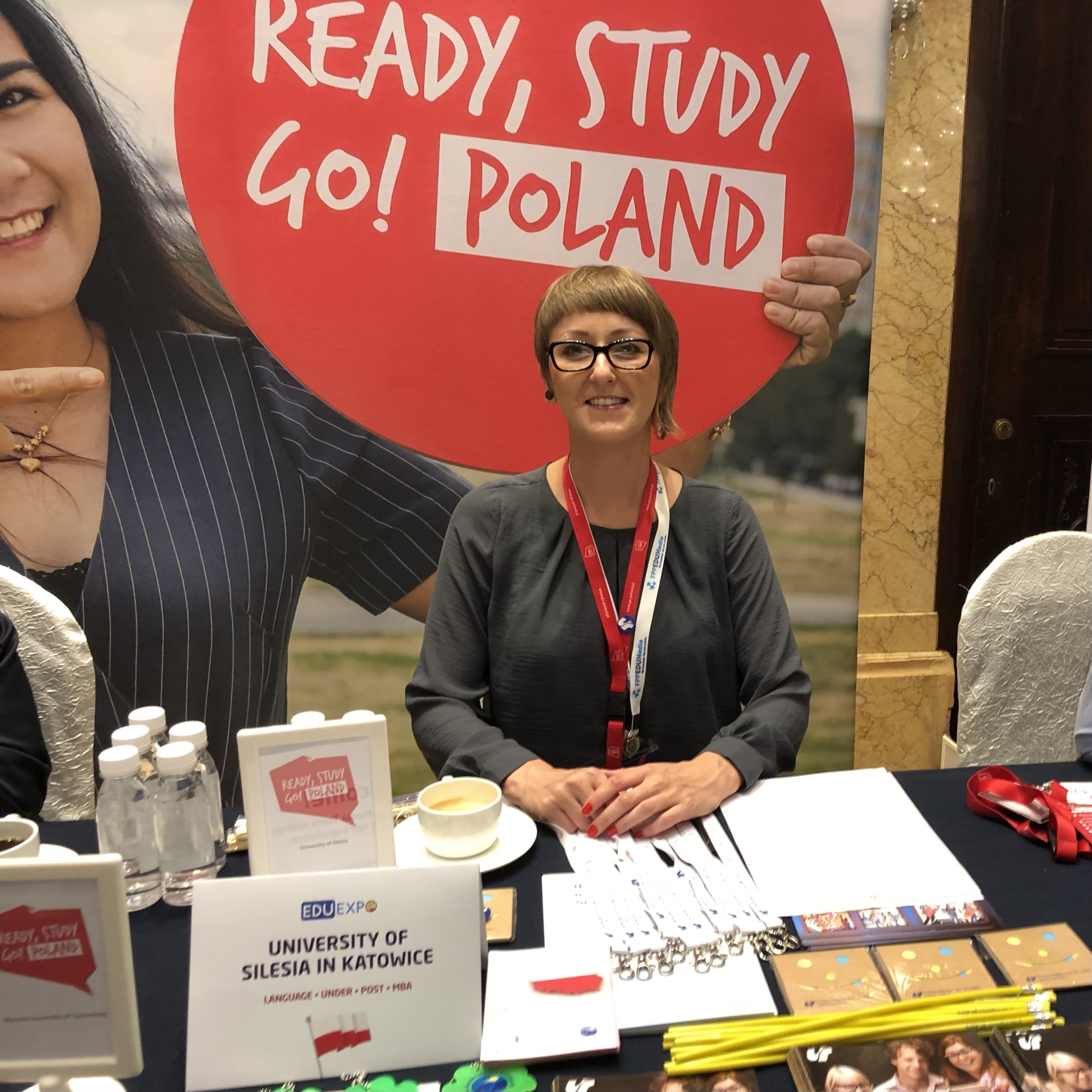Stoisko na targach edukacyjnych. Za stołem: Joanna Laskowska, w tle napis: READY, STUDY! GO POLAND. Napis na wizytówce: University of Silesia in Katowice