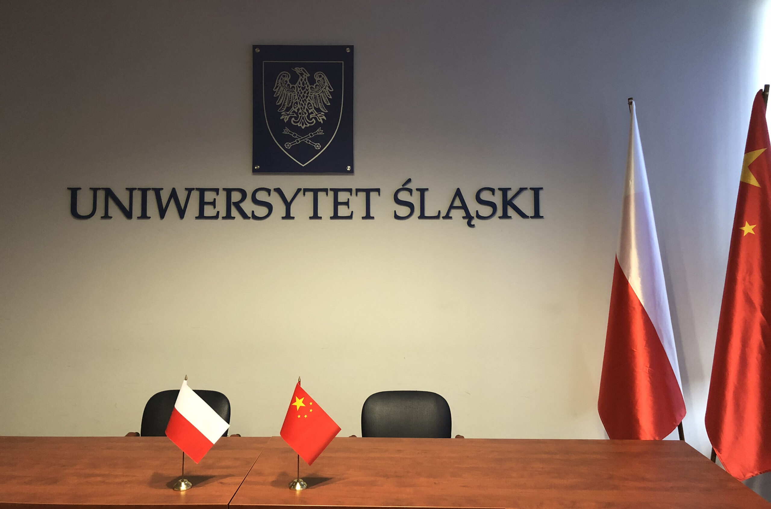Zdjęcie sali. W tle napisL Uniwersytet Śląski i herb.  Na stole i z boku flagi: Polska i  Chińska.