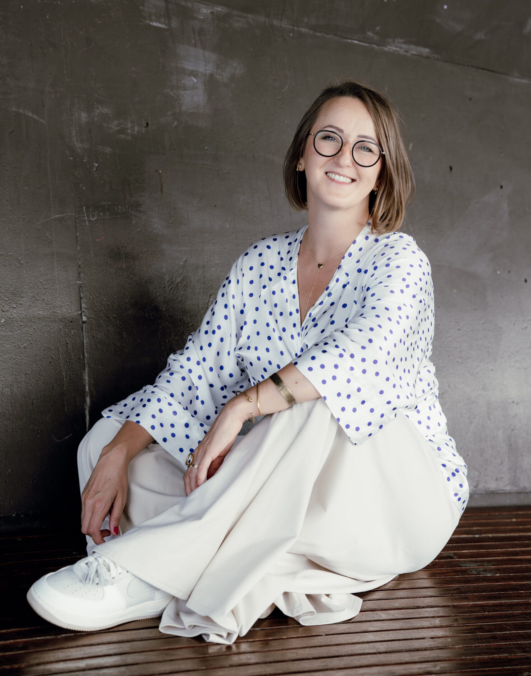 Joanna Laskowska siedzi po turecku na ławce, w tle betonowa ściana. Obrana w białką bluzkę w granatowe kropki, kremowe szerokie spodnie i beżowe buty sportowe. Uśmiecha się. Na nosie ma okulary korekcyjne